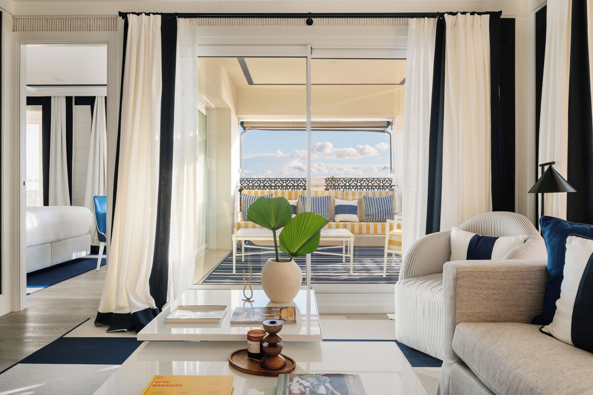 Une chambre d’hôtel avec un balcon, un canapé, une table basse agrémentée d’un vase et de bougies, et une vue sur le ciel.