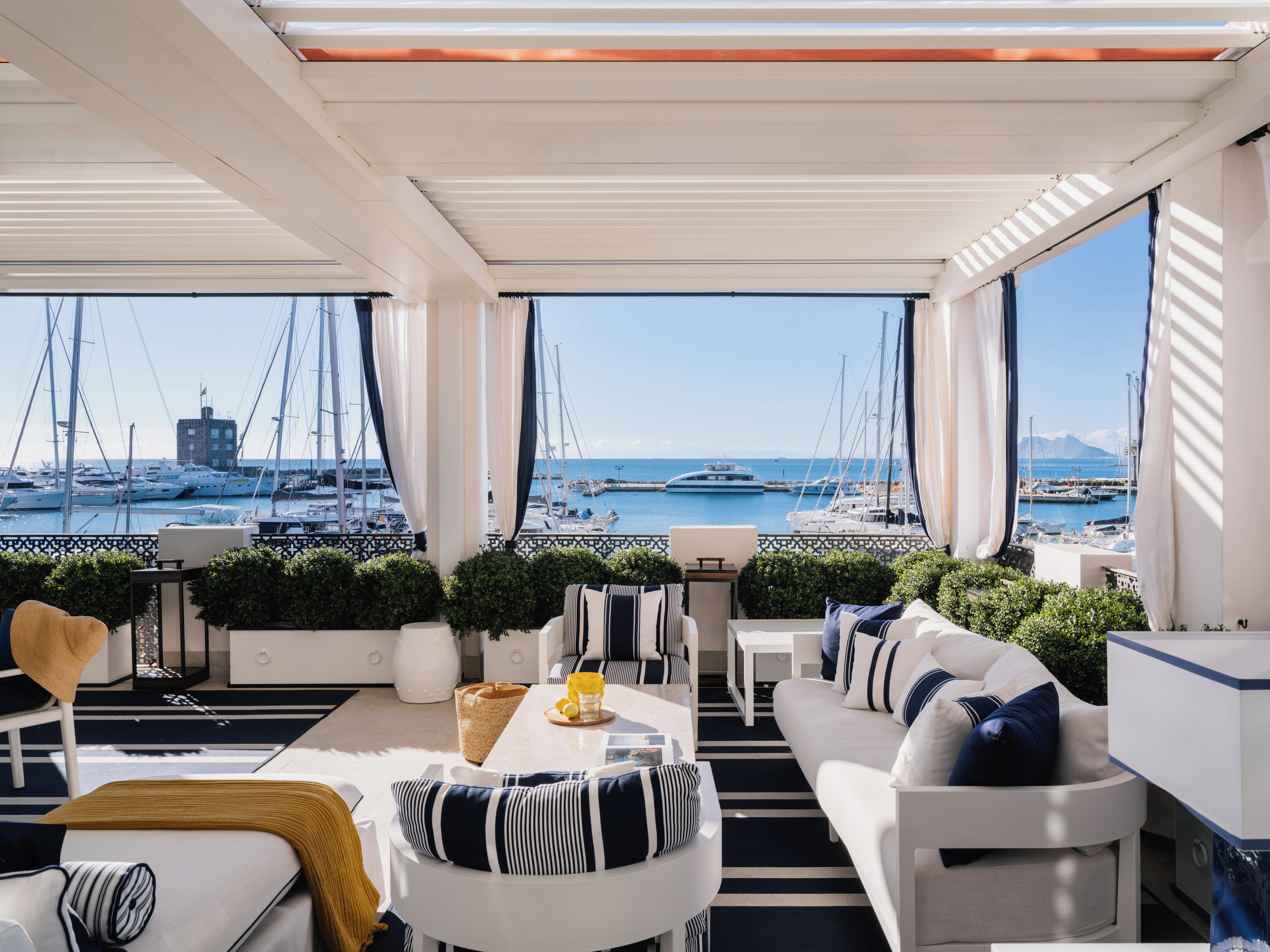 Un balcon moderne et bien éclairé, offrant une vue sur un port de plaisance et des bateaux. On y trouve des canapés blancs, une table à manger et des plantes en pot.