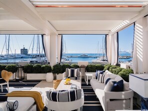 A modern, well-lit balcony with a view of a marina and boats. There are white sofas, a dining table, and potted plants. - MIM Sotogrande Member of Meliá Collection (Cadiz)