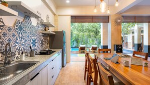 A modern kitchen with a large island, a built-in oven, and a sink. There is a dining area with wooden chairs and a table, and a view of a pool and greenery outside. - Cove Asanaya (Canggu)