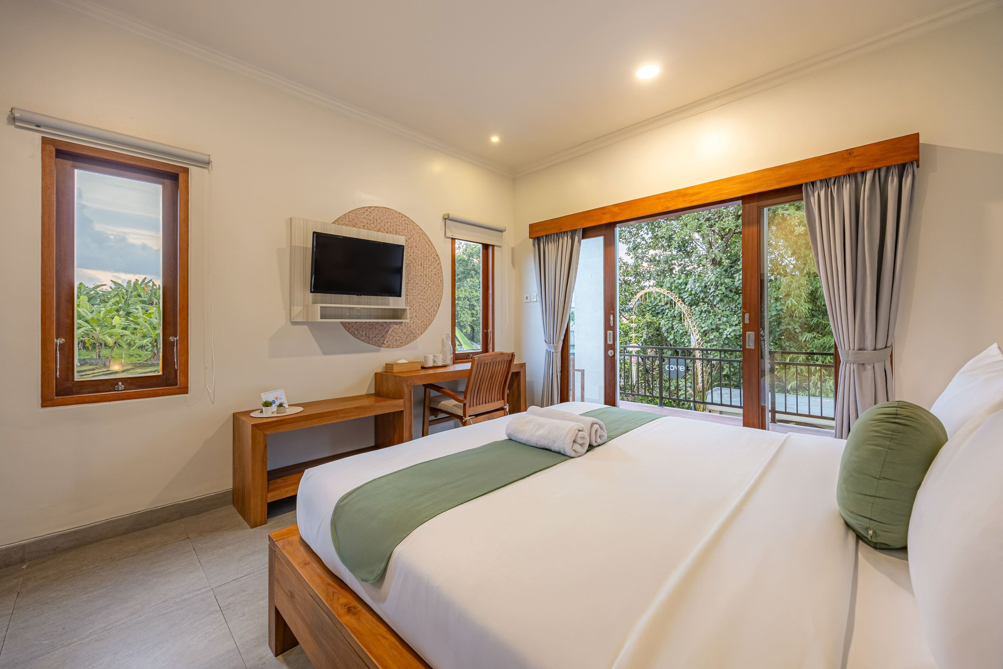 A hotel room with a bed, a desk, a television, and large windows with curtains.