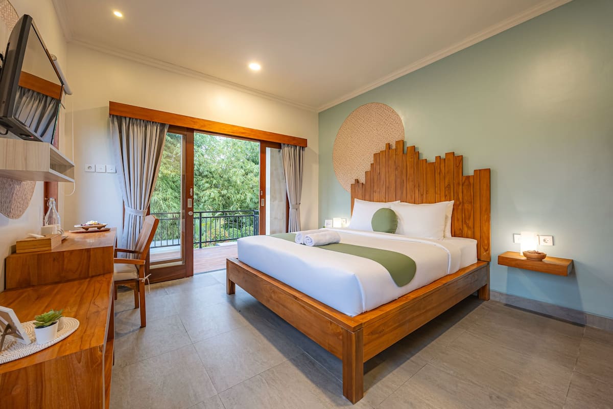 Une chambre spacieuse avec un lit en bois, un bureau avec une chaise, une petite table avec un bol, et une grande fenêtre avec des rideaux.