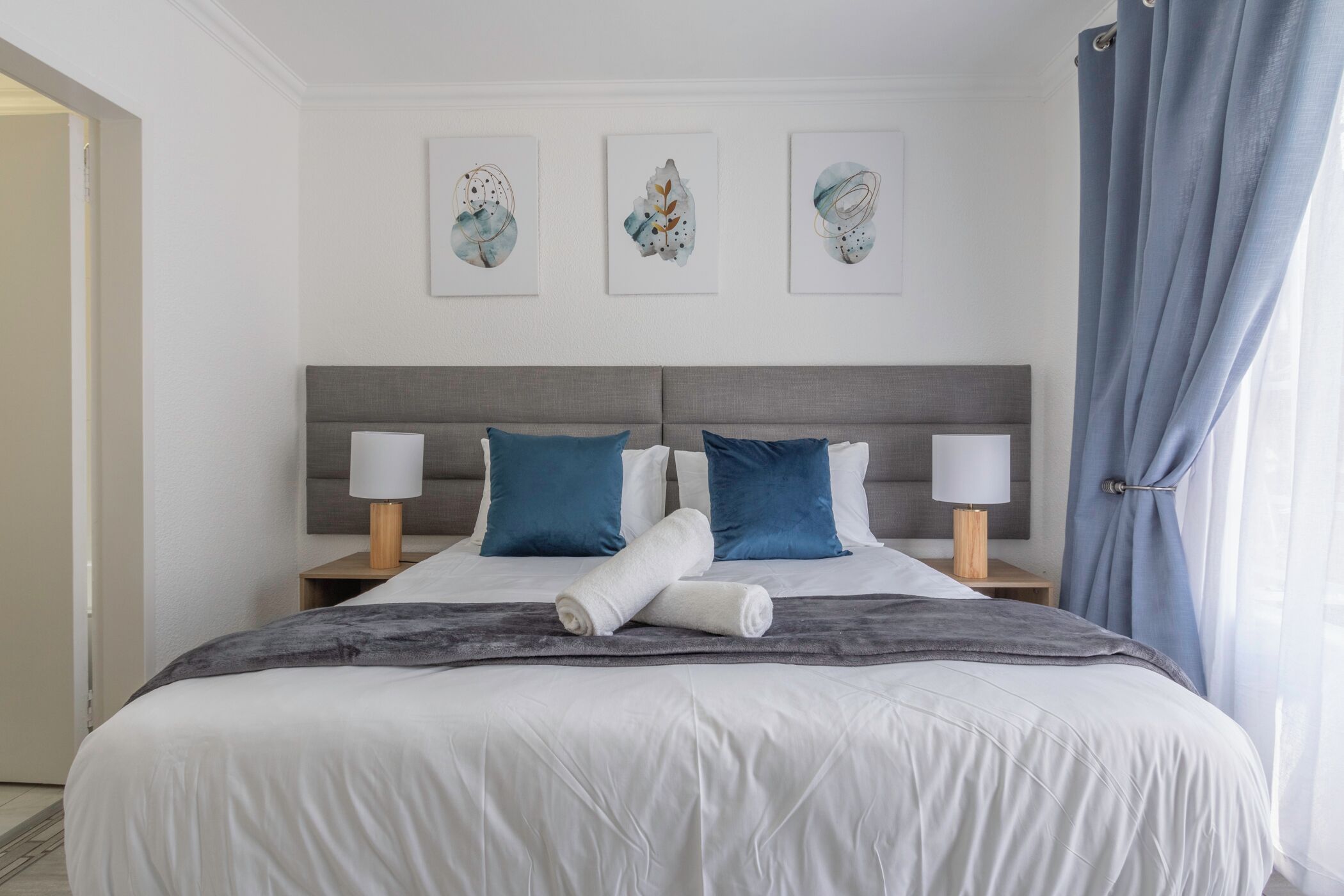 A neatly made bed with a grey headboard, white linens, and two blue pillows. There are three wall-mounted artworks above the bed.