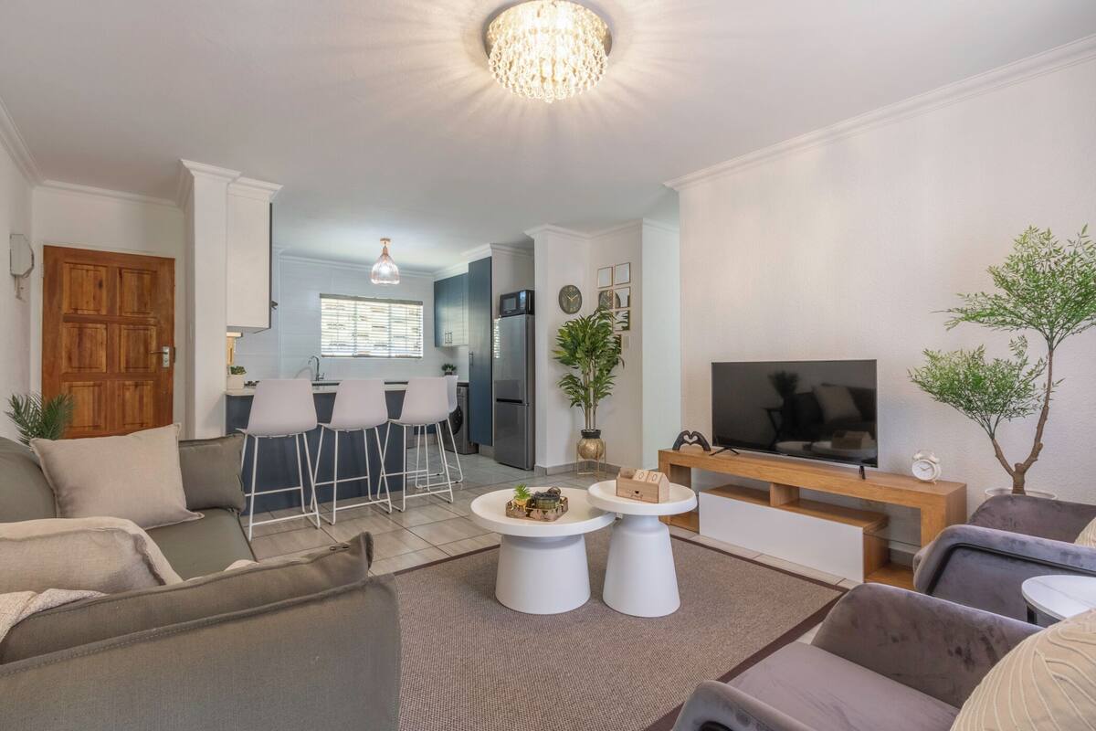 Un salon moderne avec un canapé, deux petites tables basses blanches, un téléviseur et un coin repas comprenant des chaises, avec une cuisine en arrière-plan.