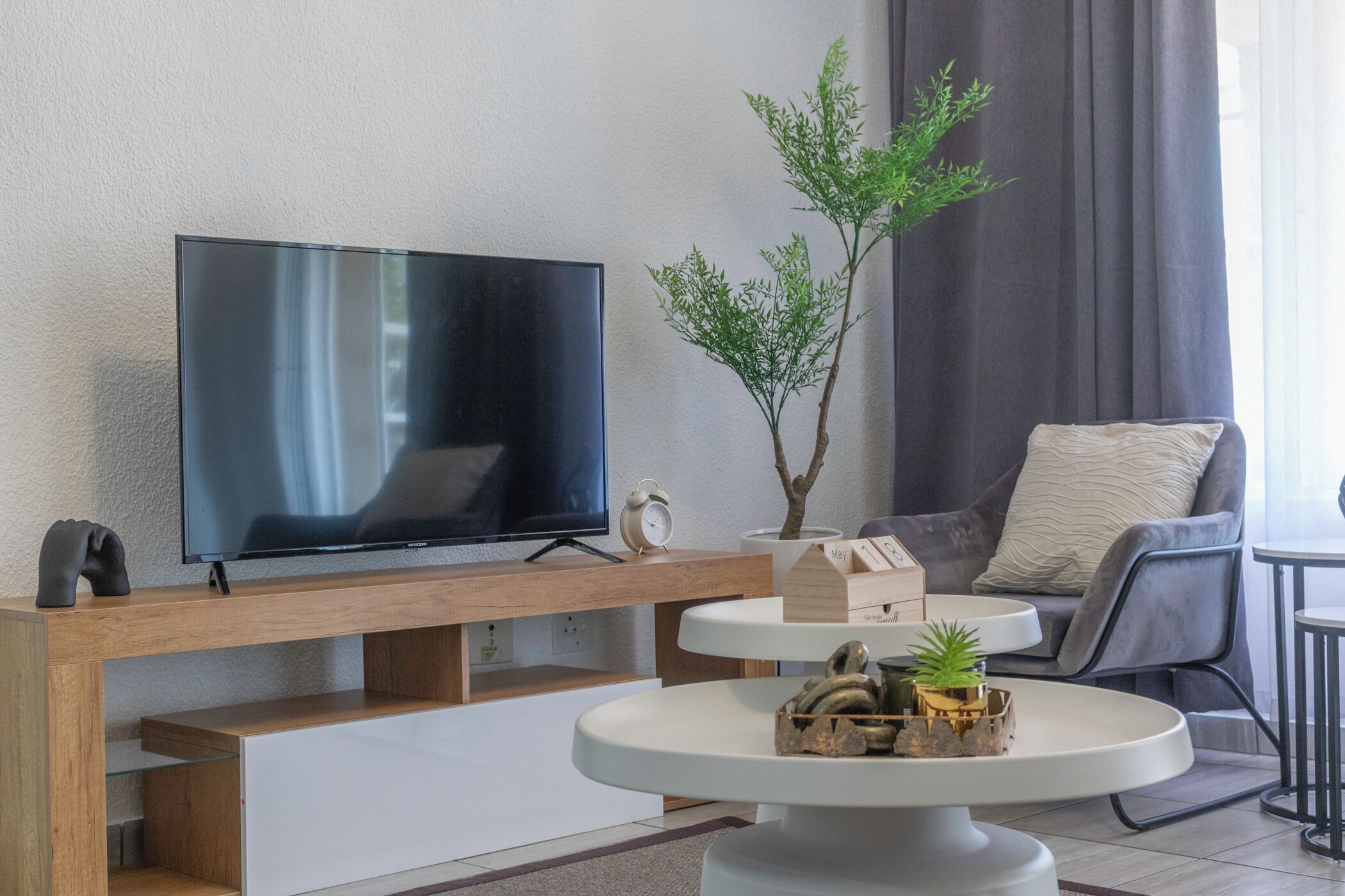 Ein modernes Wohnzimmer mit einem Flachbildfernseher auf einem Holzboden, einem kleinen Tisch mit Dekorationen, einem grauen Sofa und einer Topfpflanze.