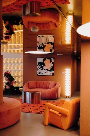 A modern interior with a sectional sofa, a round ottoman, and a wall-mounted light fixture. - LILYBETH HOTEL PRATUNAM (Bangkok)