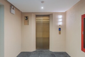 A hallway with a closed elevator, a wall-mounted light fixture, and a door with a red frame. - LILYBETH HOTEL PRATUNAM (Bangkok)