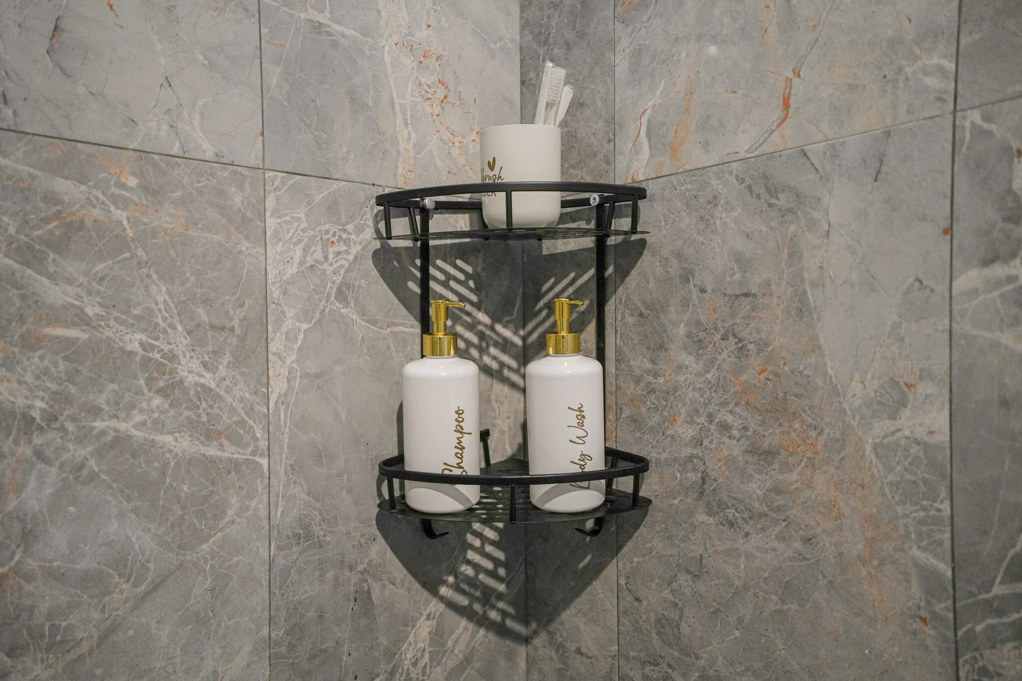 Baño con pared de mármol, estante metálico negro con un cepillo de dientes, una taza blanca y dos botellas de champú y gel de baño.