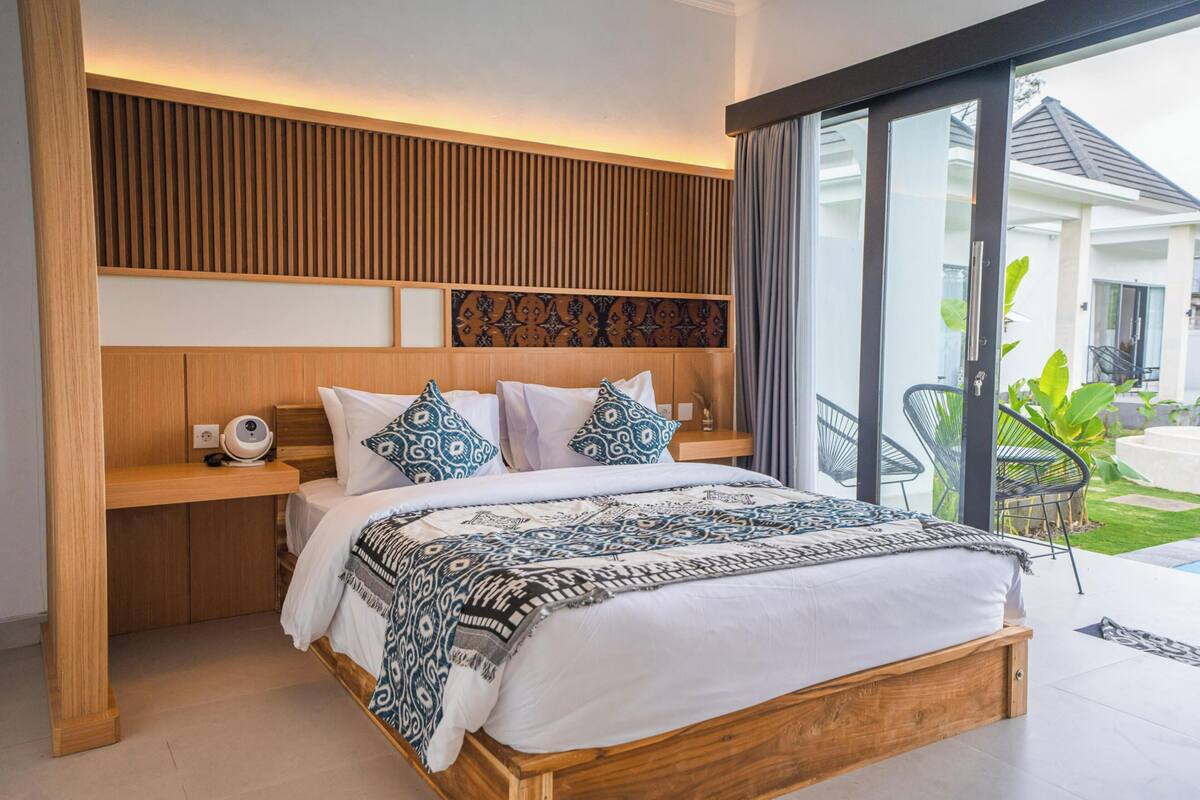 A modern bedroom with a wooden headboard, a large bed with patterned bedding, a bedside table, and a view of a garden through a sliding glass door.