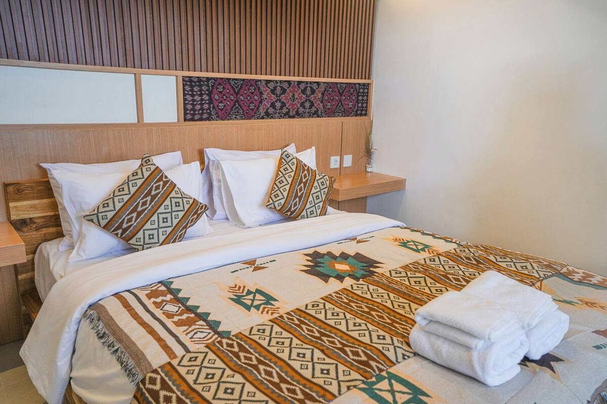 A neatly made bed with patterned bedding and pillows, a wooden headboard, and a bedside table with a vase.