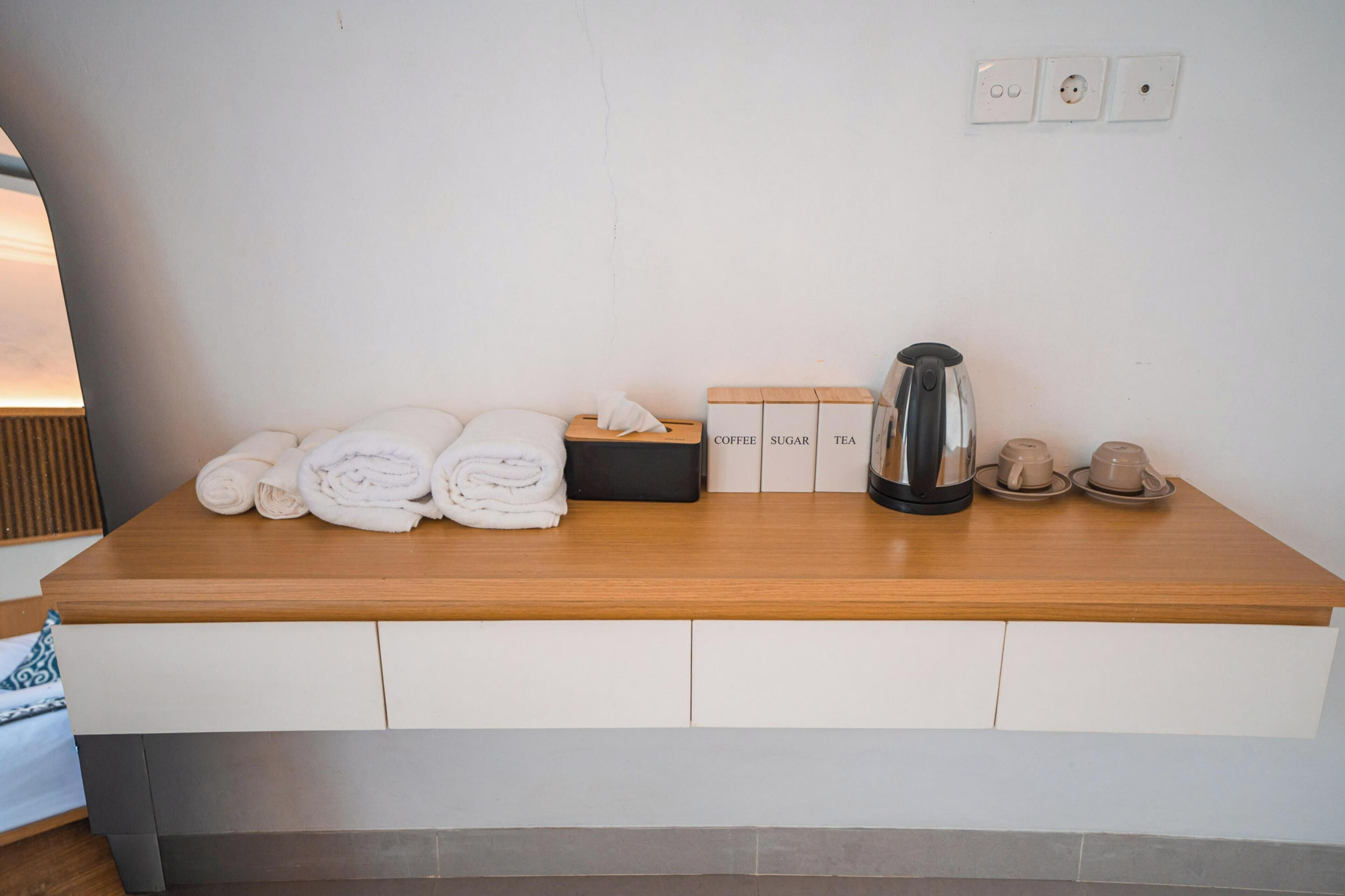 Habitación de hotel ordenada con un mostrador de madera, toallas dobladas con cuidado, una estación de café y una de té.