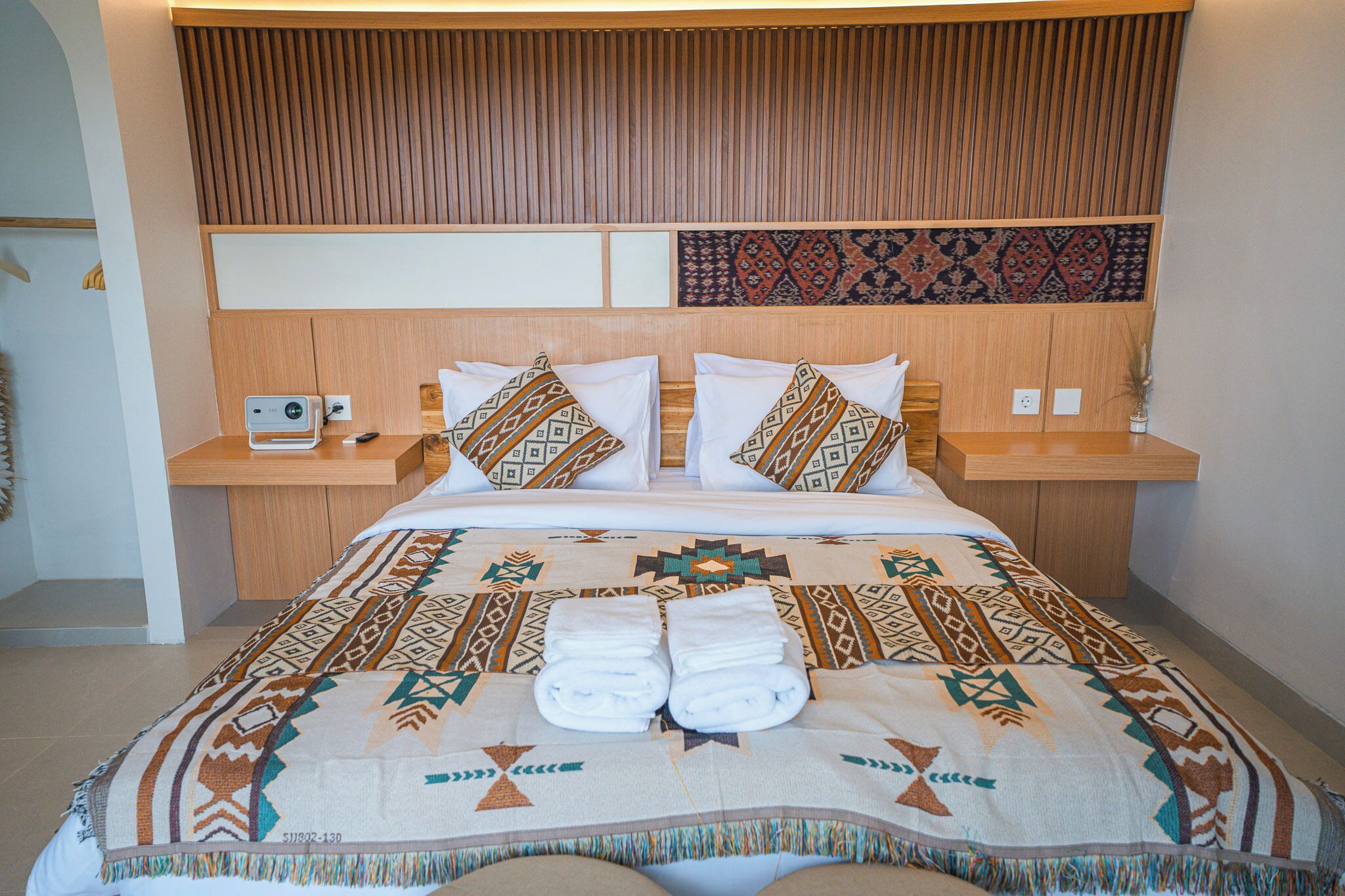 A hotel room with a large bed, wooden headboard, patterned pillows, and a neatly folded towel set.