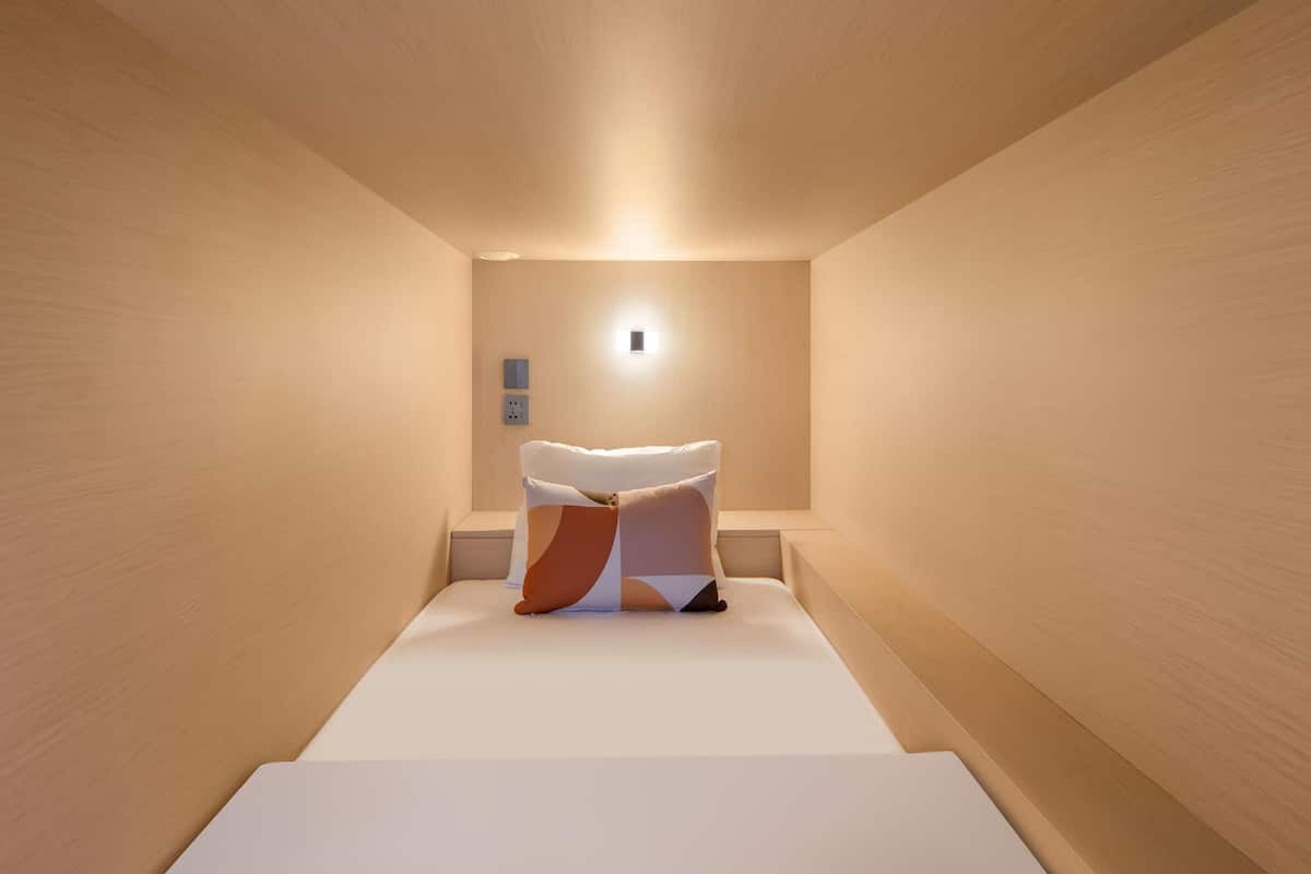 Een minimalistische hotelkamer met een bed, centraal geplaatst op een verhoging, een klein nachtkastje en een wandlamp.