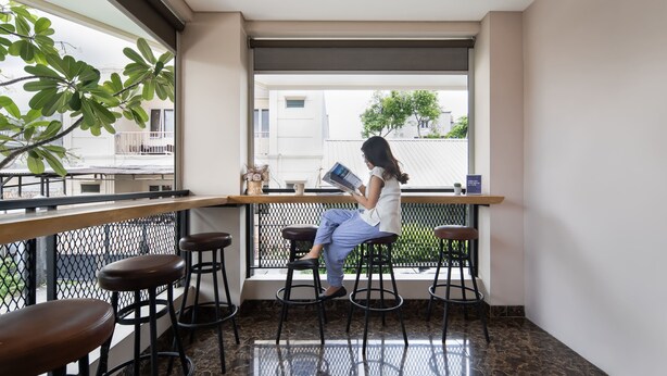 Una mujer sentada en un taburete de bar junto a una ventana, leyendo un libro. Hay tres taburetes y una mesa con una planta y una taza.