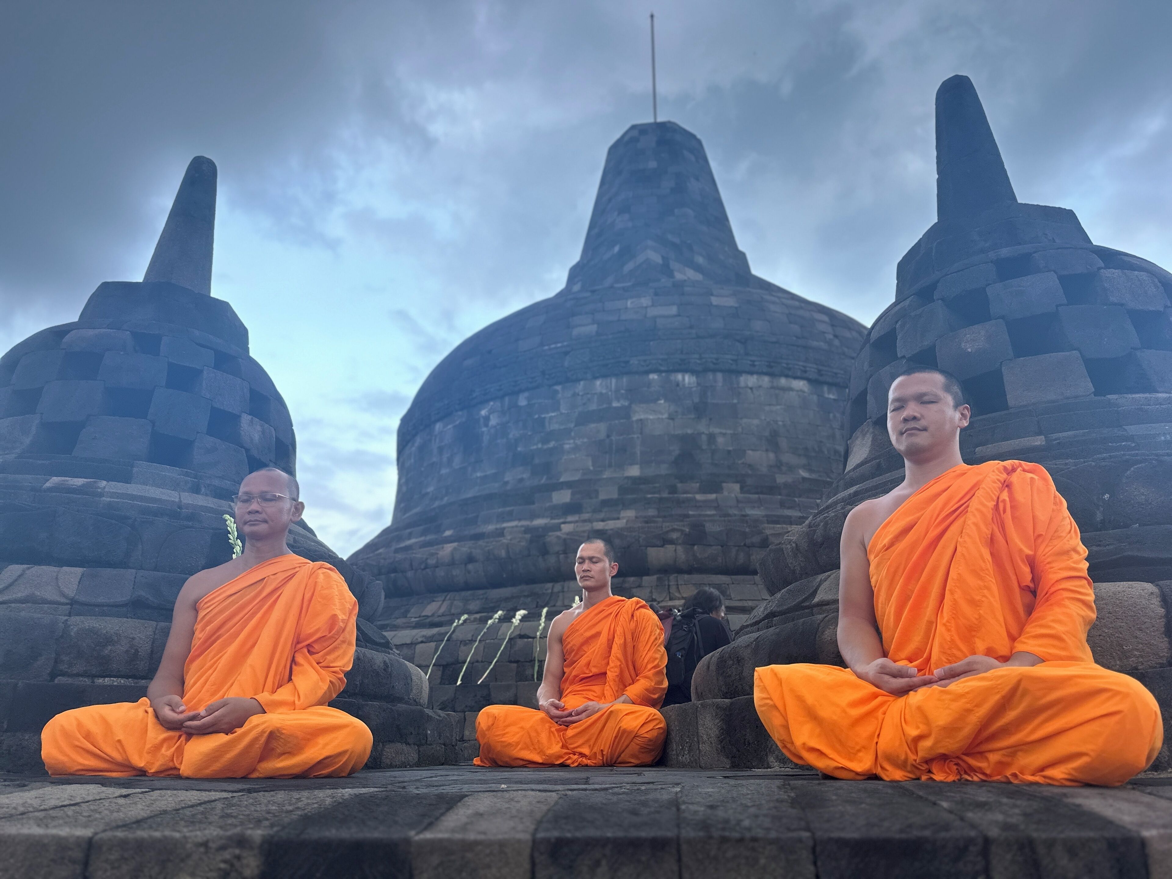 Tres personas vestidas con túnicas anaranjadas sentadas en posición meditativa frente a una gran estructura de piedra antigua con techo puntiagudo.