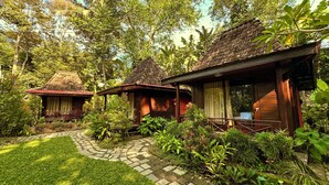 A traditional wooden house with a tiled roof, surrounded by lush greenery and a stone pathway.