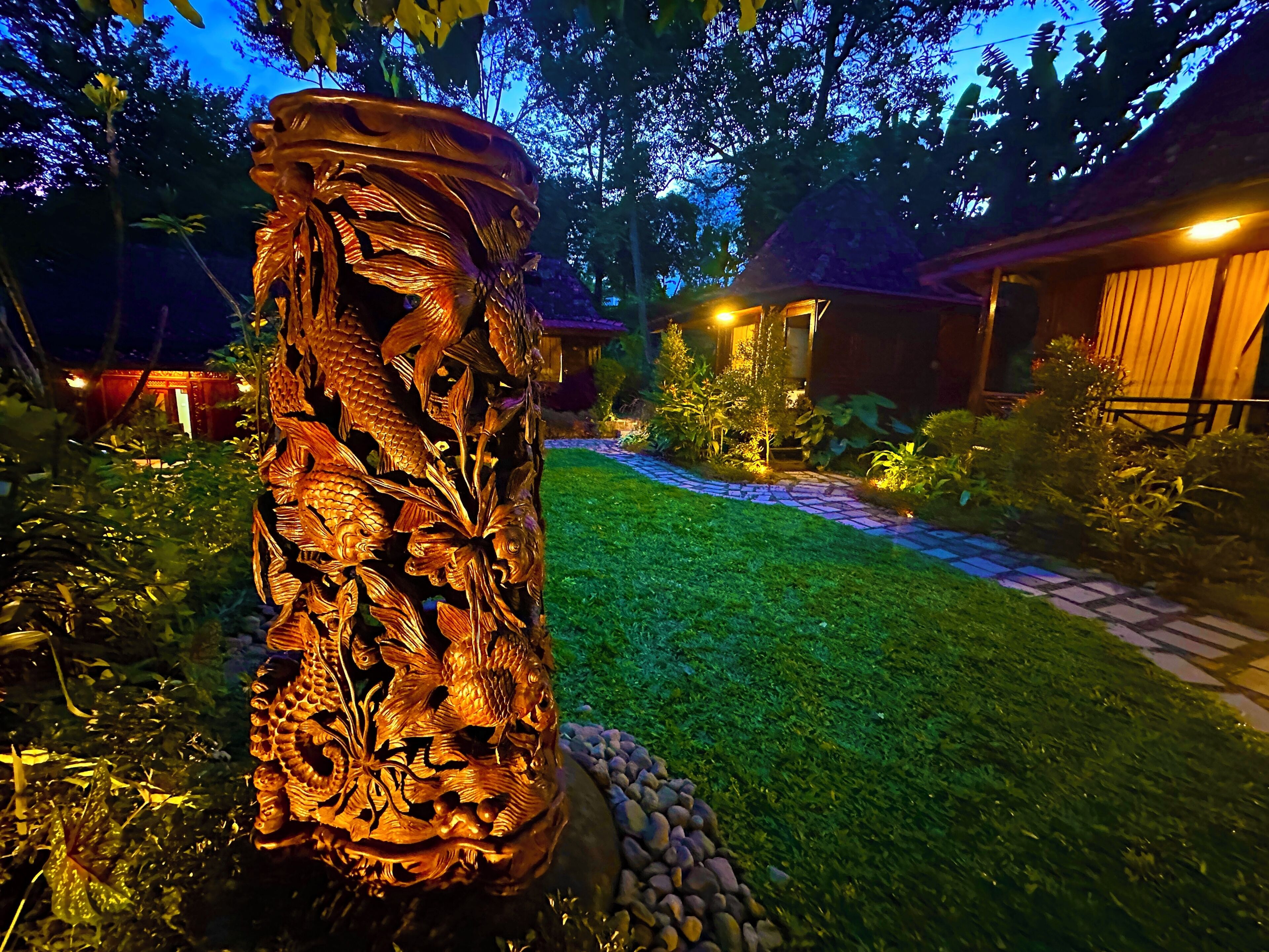 Um jardim com uma grande escultura de madeira ricamente detalhada com peixes e folhas, um caminho de pedra e um edifício com telhado de palha ao fundo.