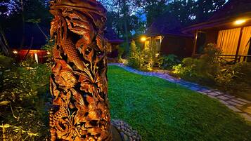 Um jardim com uma grande escultura de madeira ricamente detalhada com peixes e folhas, um caminho de pedra e um edifício com telhado de palha ao fundo.