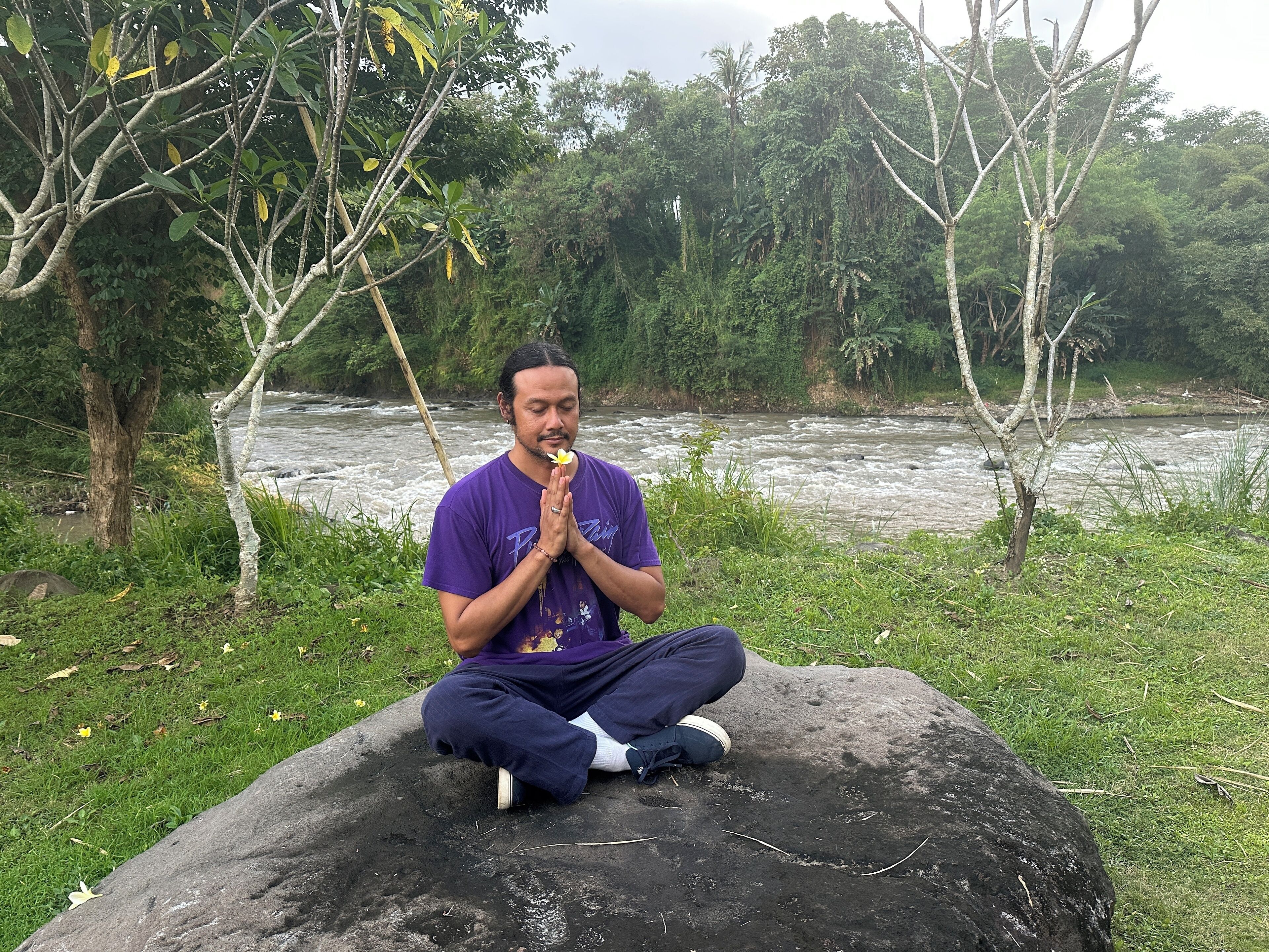 A person sitting on a rock by a river, surrounded by greenery.