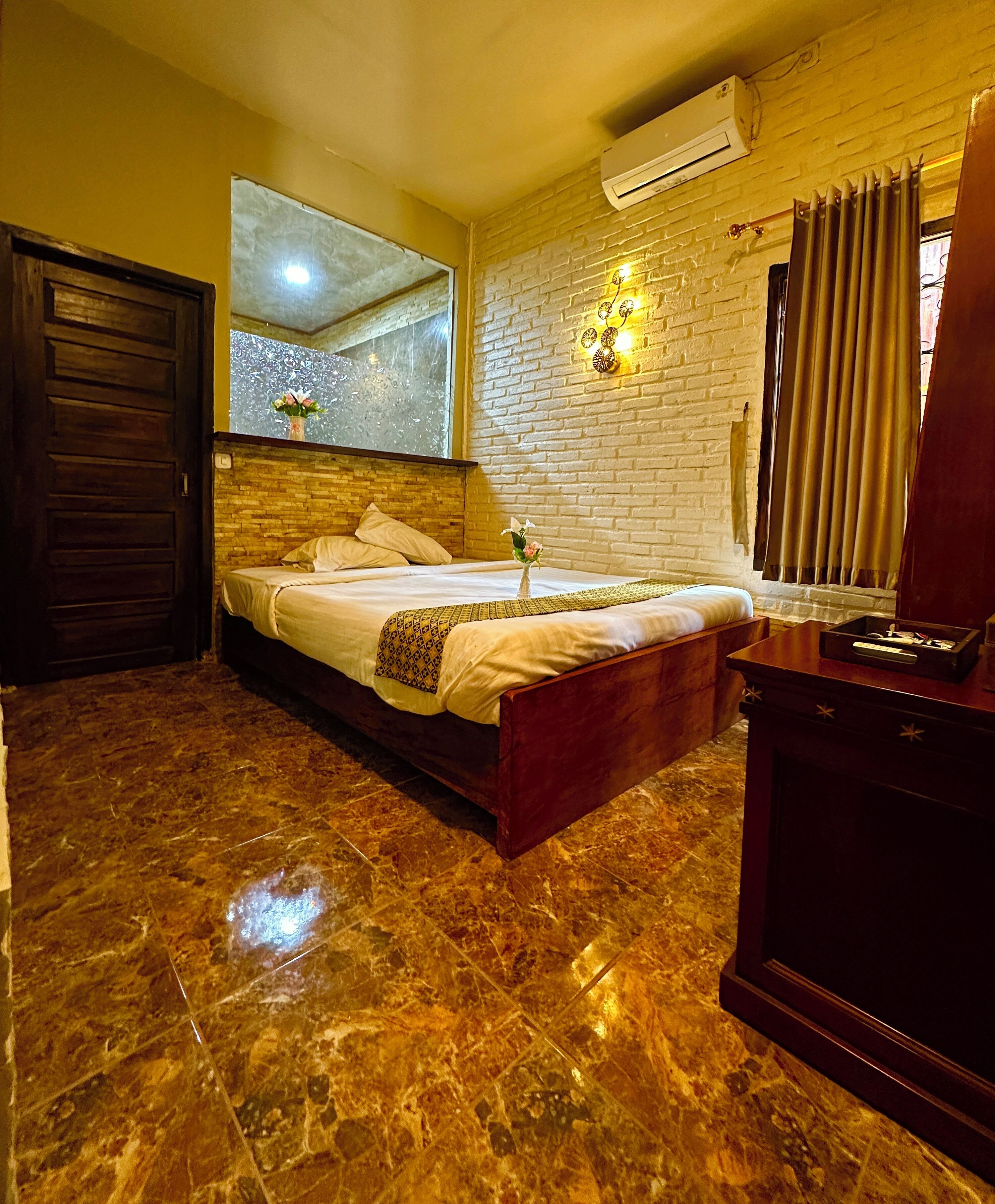 Un dormitorio con cama, cómoda de madera, piso de mármol, pared de ladrillo y equipo de aire acondicionado.