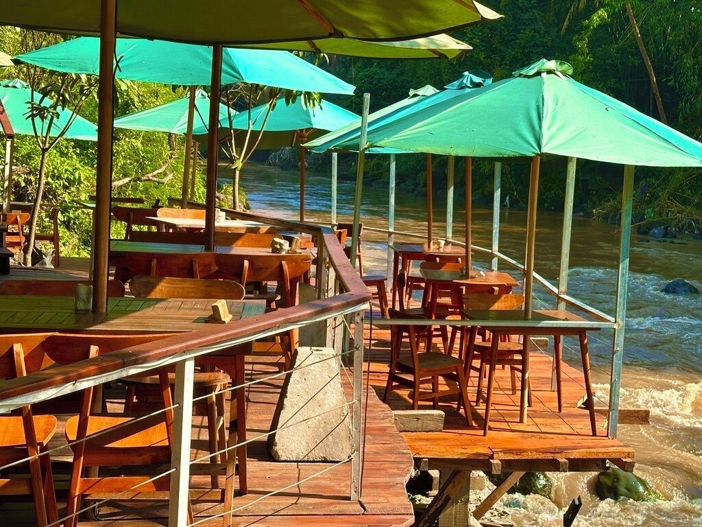 Terraza de madera con mesas y sillas, rodeada de sombrillas verdes, con vista al río.