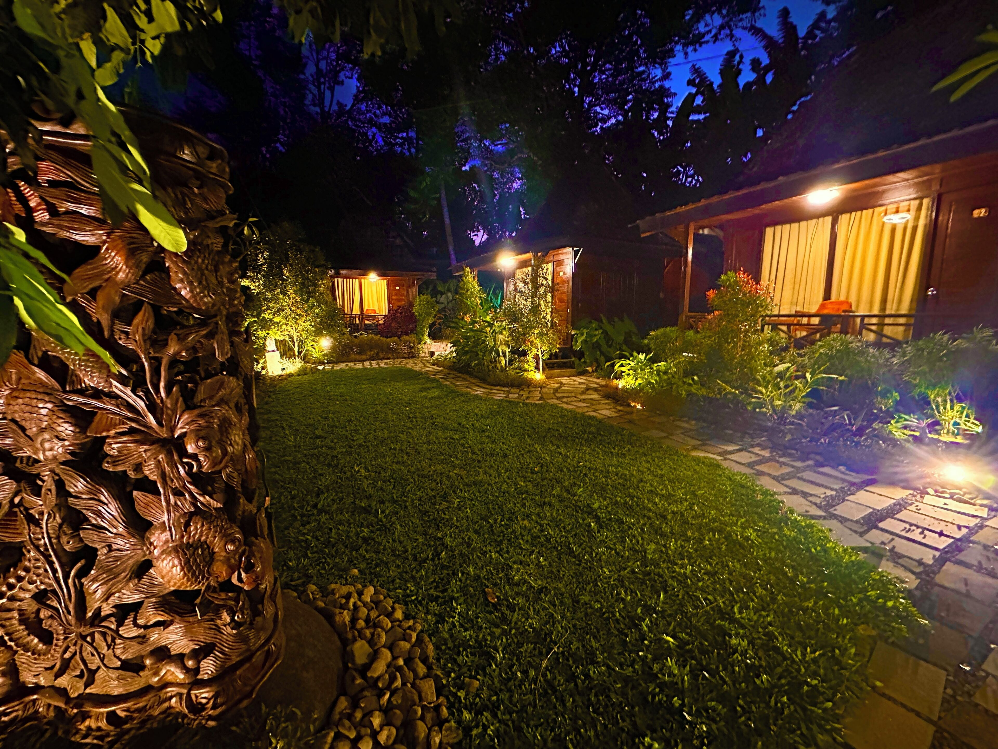 A nighttime garden with a stone pathway, a wooden sculpture, and two lit buildings with curtains.