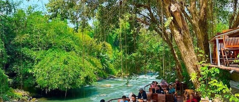 Ein Flussrestaurant mit Außenbereich, das einen Fluss mit Blick auf das sanft fließende Wasser bietet, umgeben von üppiger Vegetation.