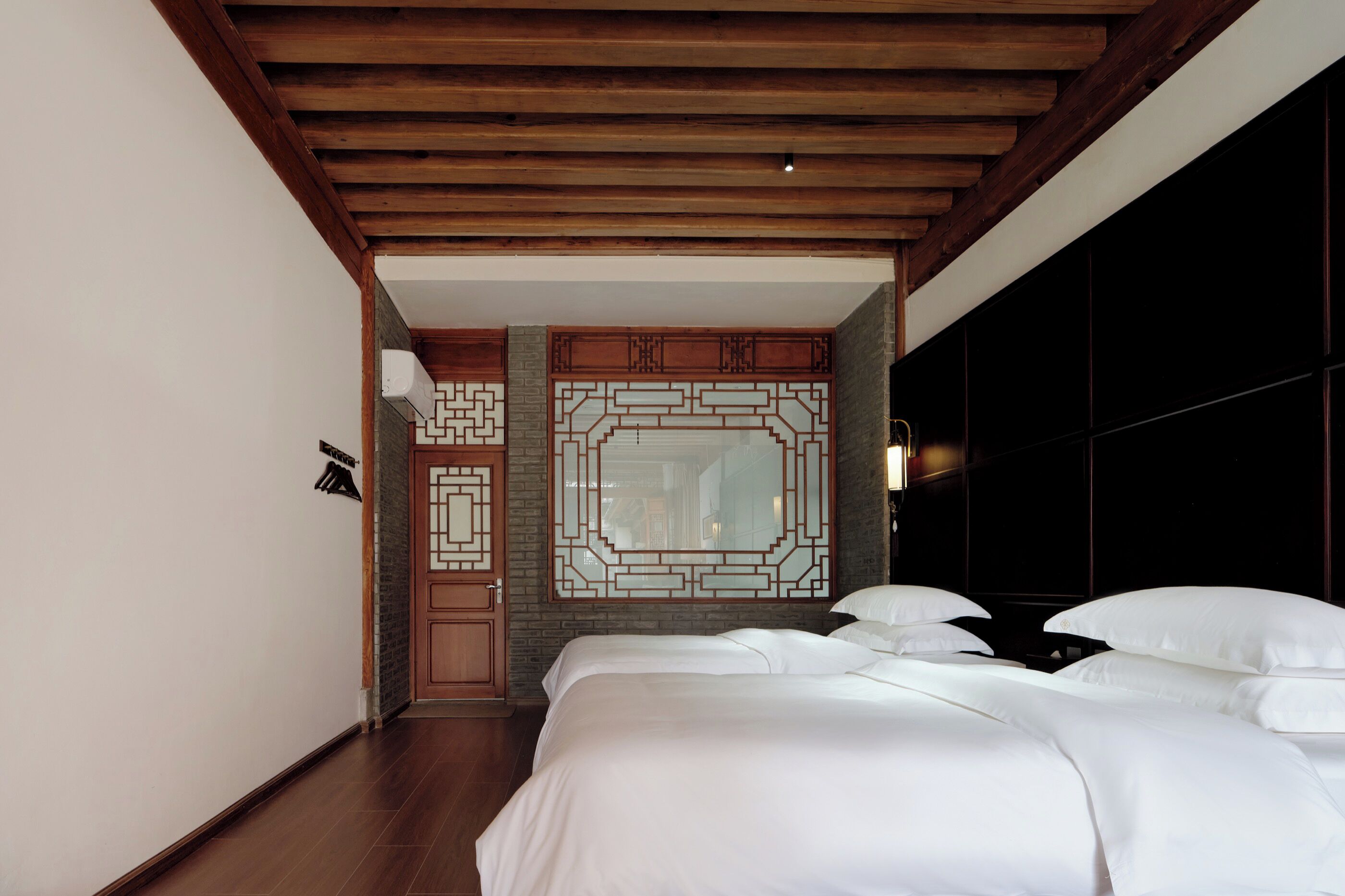 A hotel room with a wooden ceiling, a large bed with white linens, a decorative wall panel, and a dark room with a large window.