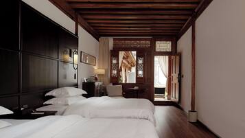 A hotel room with a wooden ceiling, a bed with white bedding, a desk with a lamp, and a view through a door to an outdoor area.