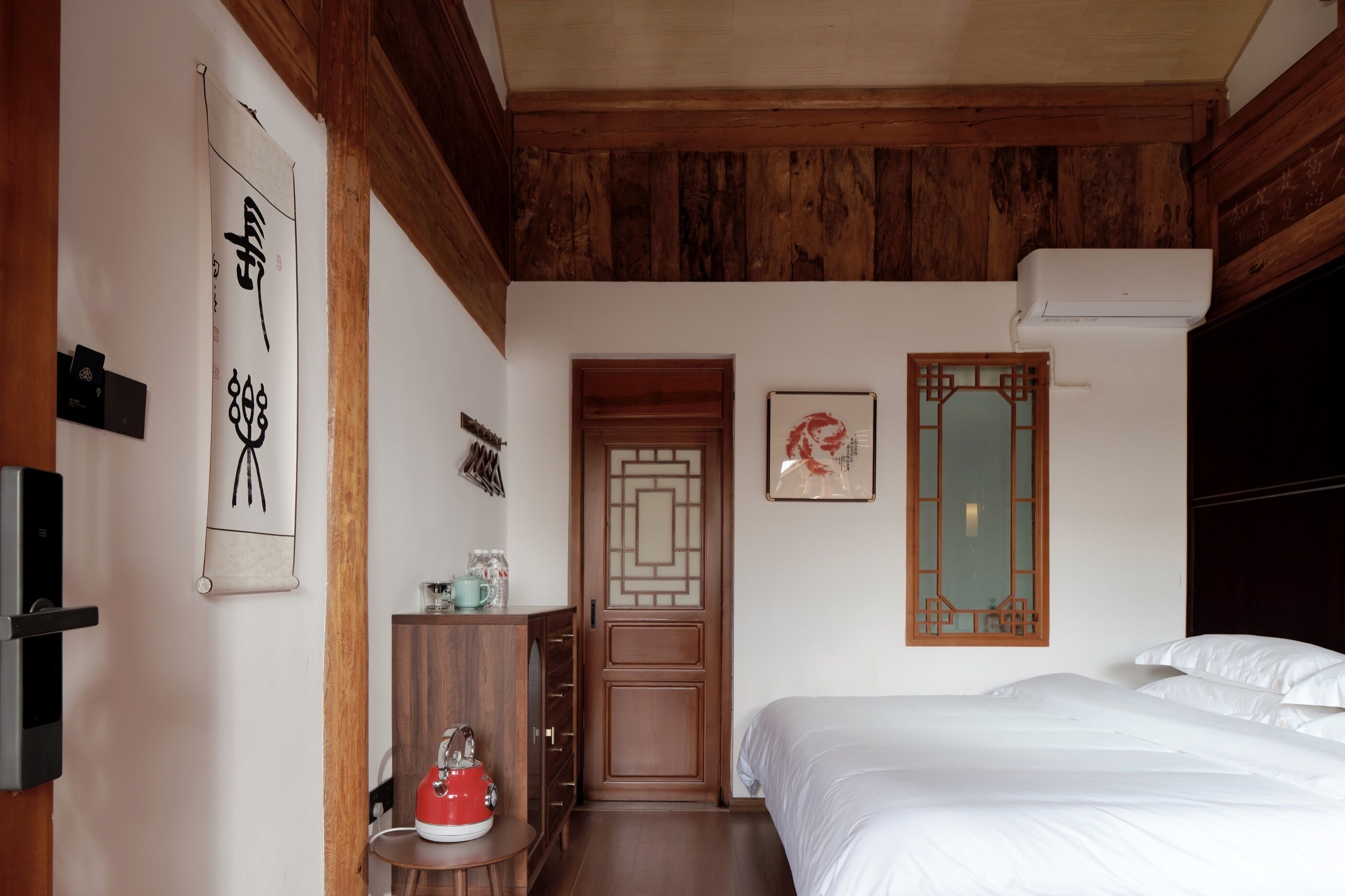 A bedroom with a wooden ceiling, a bed with white bedding, a wooden wardrobe, a small table with a kettle, and a door with traditional Chinese design.
