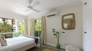A bedroom with a bed, a ceiling fan, an air conditioning unit, a mirror, a window with curtains, a plant, and a view of greenery outside. - Top Floor Retreat – Walk to Beach & Town (Port Douglas)