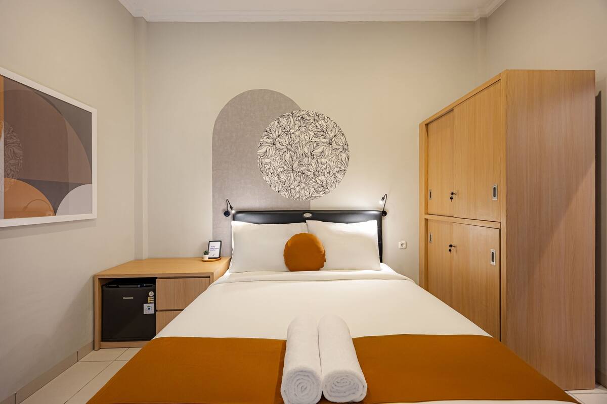 A hotel room with a bed, a wooden wardrobe, a desk with a phone, and a wall art piece.