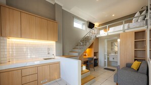 A modern kitchen with wooden cabinets, a white countertop, and a built-in oven. There is a staircase leading to a loft bedroom with a bed and a TV. - Cove Nuansa Home (Jimbaran)