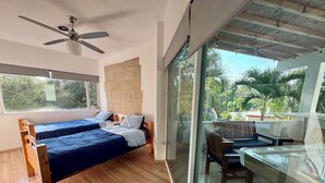 Une chambre avec deux lits en bois, un ventilateur de plafond, une grande fenêtre et un balcon équipé d’une table et d’une chaise.