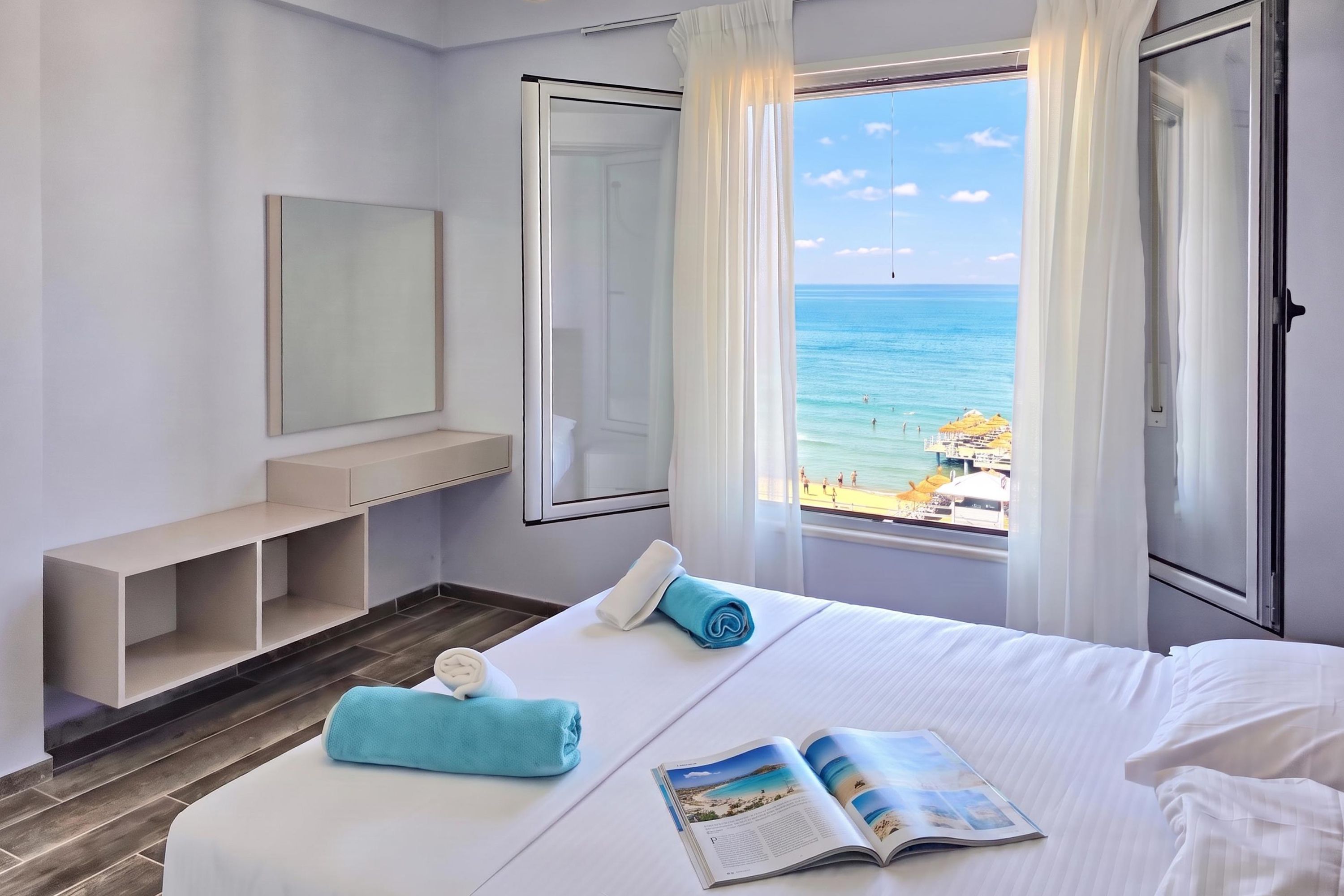 A hotel room with a bed, a bedside table, a mirror, and a window with a view of the beach.