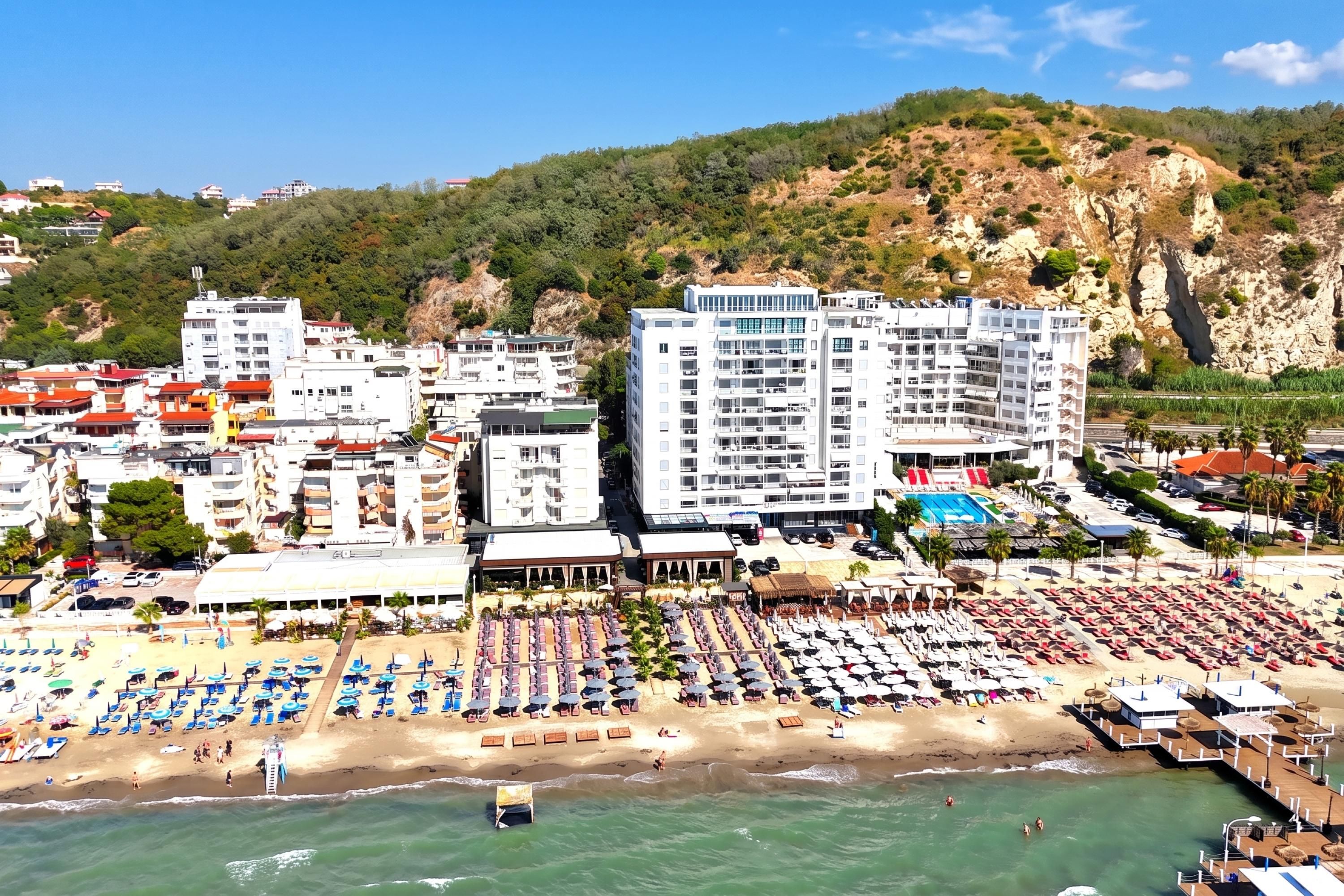 Un hotel costero con varios pisos, una playa con numerosas tumbonas y sombrillas, y una zona de piscina.