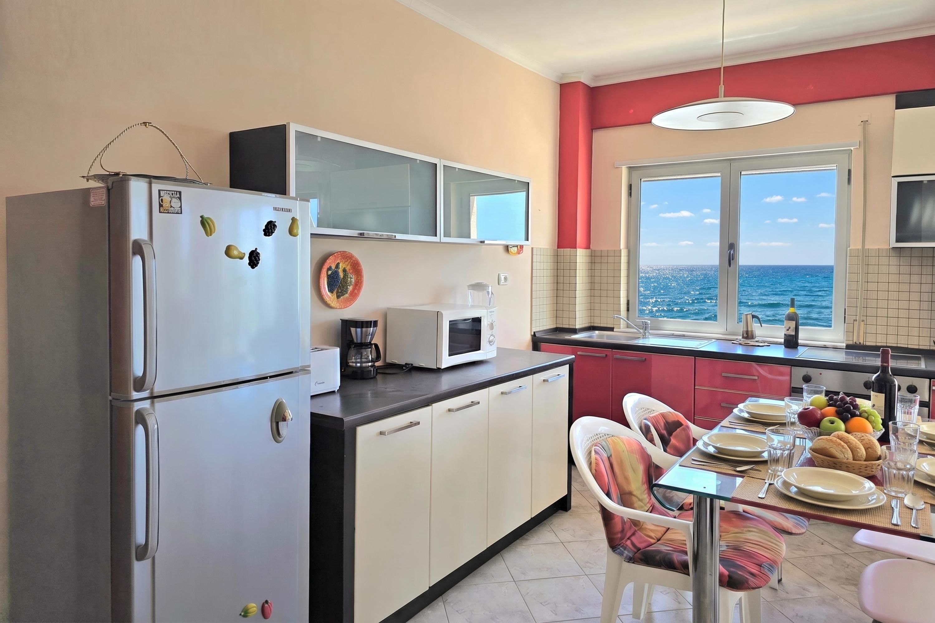 Eine moderne Küche mit Kühlschrank, Mikrowelle und Kaffeemaschine. Es gibt einen Essbereich mit Tisch für zwei Personen mit Blick auf eine schöne Meerlandschaft.