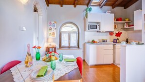A dining area with a table set for two, a kitchen with white cabinets, and a window with a view of greenery. - Le Fontane With View Lake, Gardone Riviera, Italy (Gardone Riviera)