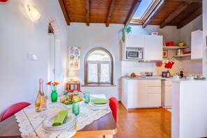 A dining area with a table set for two, a kitchen with white cabinets, and a window with a view of greenery. - Le Fontane With View Lake, Gardone Riviera, Italy (Gardone Riviera)