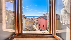 A window with a view of a narrow street with buildings, a clear sky, and a distant landscape. - Le Fontane With View Lake, Gardone Riviera, Italy (Gardone Riviera)