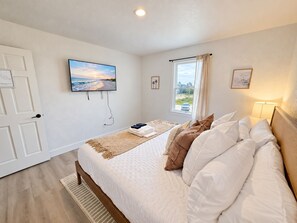 Ein Schlafzimmer mit einem Bett, einem an der Wand befestigten Fernseher, einem Fenster mit Blick auf den Strand und einer Tür.