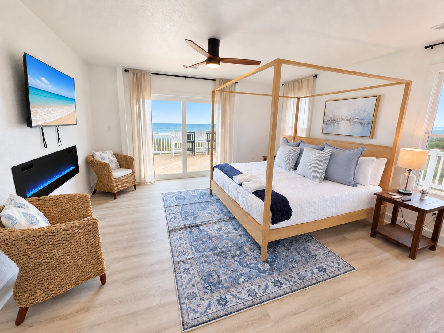 A bedroom with a four-poster bed, a television, a wicker chair, a nightstand, and a view of the beach.