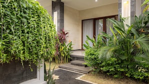 A modern house with a stone exterior, a concrete pathway leading to a wooden door, and lush greenery surrounding the property. - Cove Cubang Homestay (Kutuh)