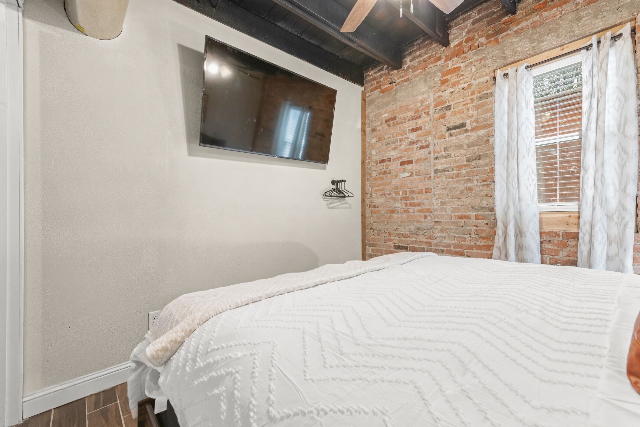Un dormitorio con pared de ladrillo, ventana con cortinas, cama con cubrecama estampado y televisor montado en la pared.
