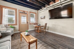 A living room with a sofa, a coffee table, a dining table with chairs, a TV mounted on the wall, and a door with a window. - Texas Cactus (Baird)