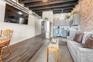 Un salon moderne avec une table à manger en bois, un canapé et un coin cuisine doté de meubles gris et d’un réfrigérateur en acier inoxydable.