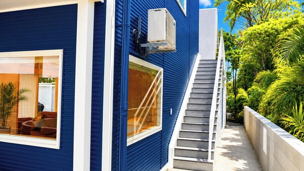 Une maison bleue avec un escalier menant à un balcon, entourée d’une végétation luxuriante.