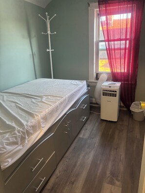A bedroom with a bed, a white heater, a window with red curtains, and a coat rack.