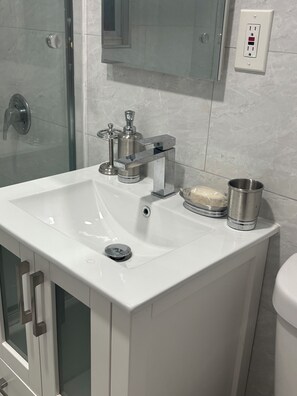 A bathroom with a white sink, a glass shower enclosure, a mirror, and a wall-mounted electrical outlet.