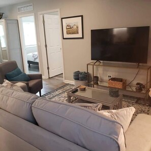 A living room with a sofa, a coffee table, a TV, and a door leading to another room. - Newly Remodeled 2BR 2BA Home  Close to Barrington (Barrington)